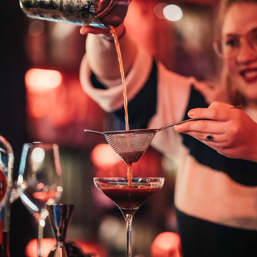 A barista is pouring a drink from a shaker through a sieve into a cocktail glass